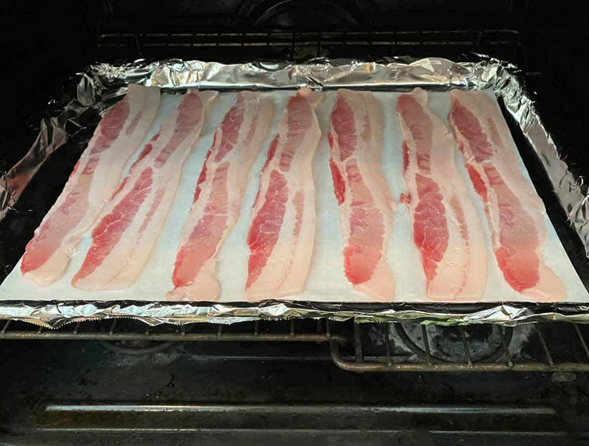 Uncooked bacon strips on a parchment and foil lined baking sheet, going into a cold oven.