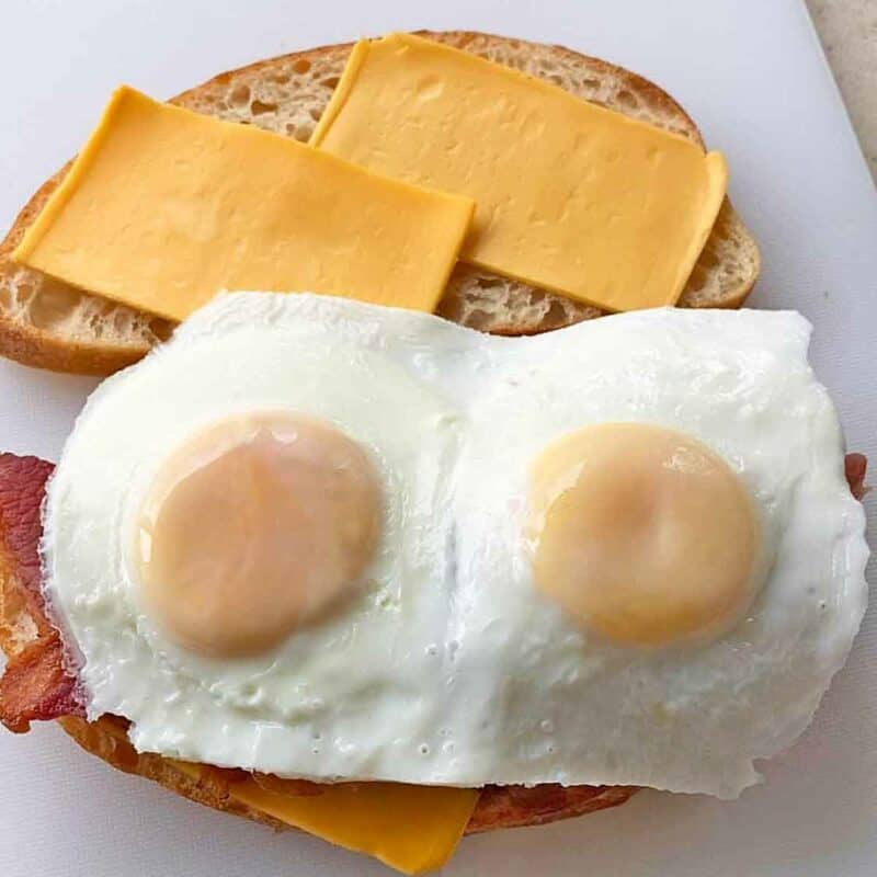 Two basted eggs placed on one piece of bread with bacon to build a sandwich.