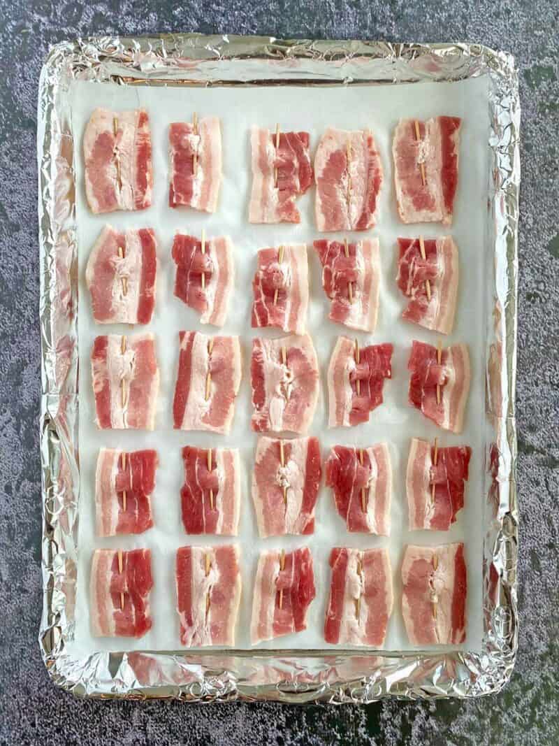 Two dozen uncooked bacon pieces on a parchment and foil lined baking sheet.
