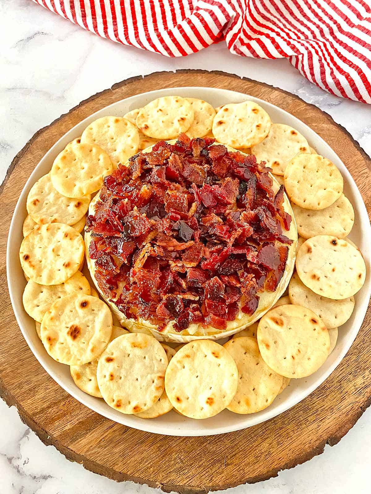 Bacon topped Brie Cheese surrounded by crackers on a serving plate.