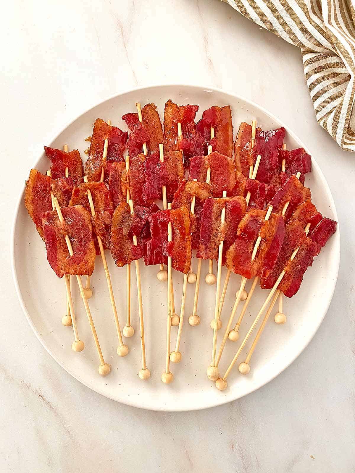 A serving plate with 24 bacon lollipops appetizers on wooden sticks.