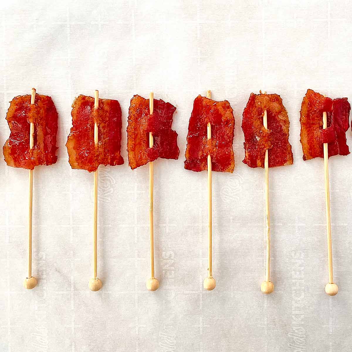 Six homemade candied bacon lollipops on wooden sticks on parchment paper.