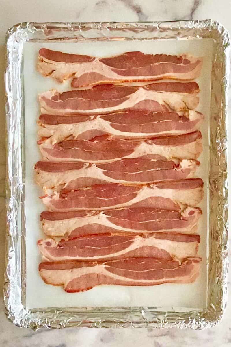 A foil and parchment lined baking sheet with uncooked bacon strips.