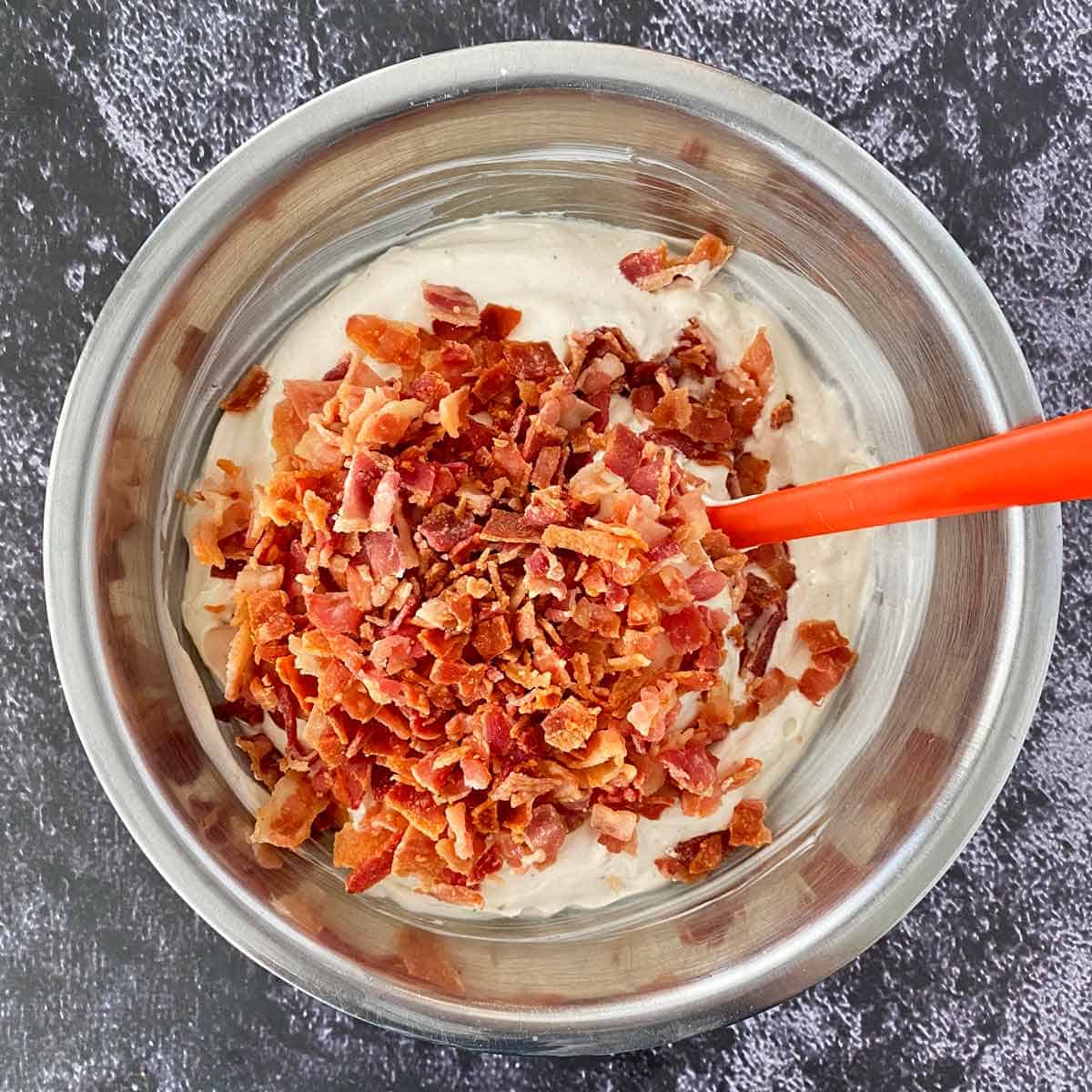 Bacon bits in a stainless steel mixing bowl with dip and a red spatula.