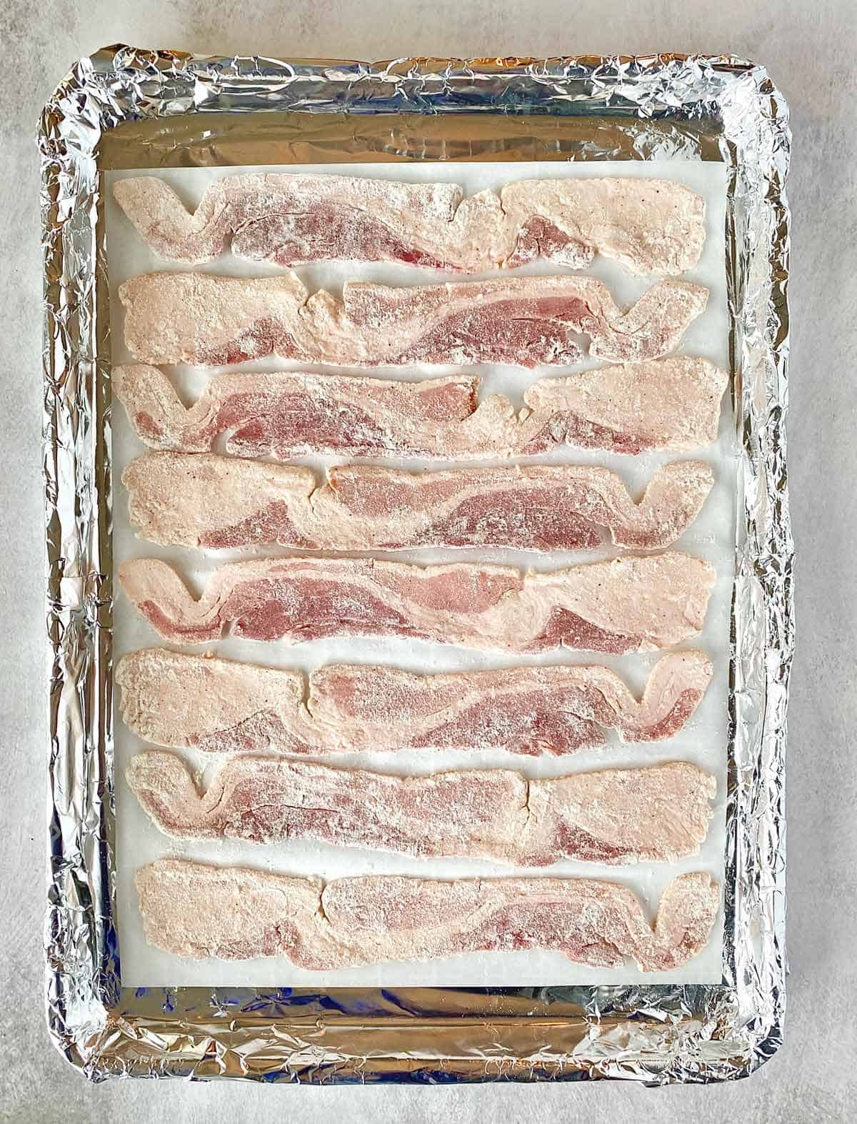 Eight slices of flour coated bacon arranged on a baking sheet.