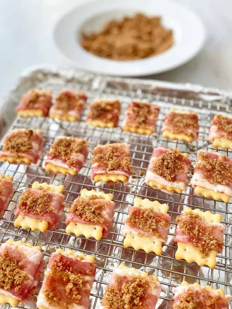 Bacon wrapped crackers sprinkled with brown sugar on a baking rack. A small bowl of brown sugar, cayenne pepper and black pepper is in the background.