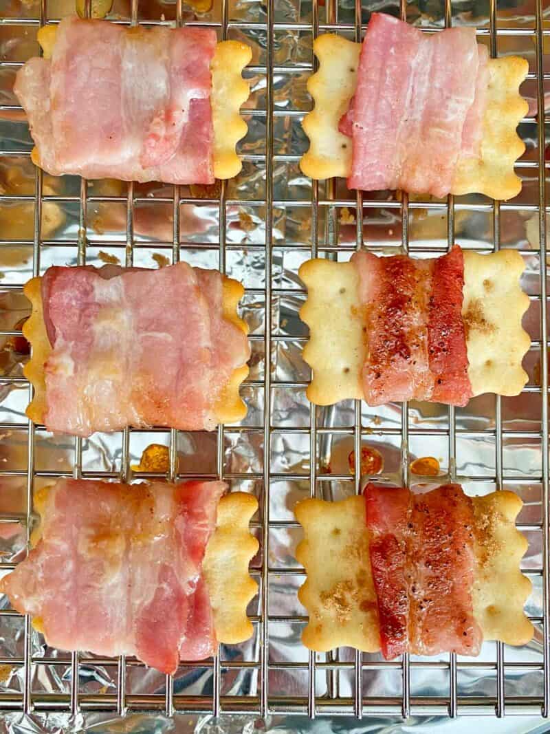 Six partially cooked bacon wrapped crackers on a rack on a foil-lined baking pan.